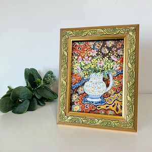 May include: A framed painting featuring a white pitcher with yellow and blue spots, filled with pink and yellow flowers. The painting has a colorful background with red, blue, and yellow patterns. The frame is gold with a green floral design.