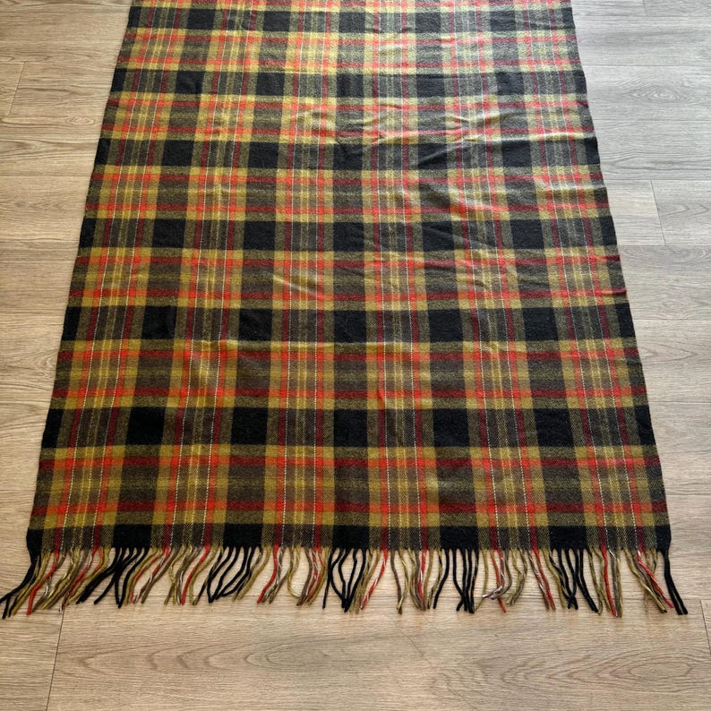 May include: A rectangular plaid blanket in a classic pattern. The blanket features a repeating pattern of black, red, and gold squares. The blanket has a fringe of black tassels along the bottom edge.