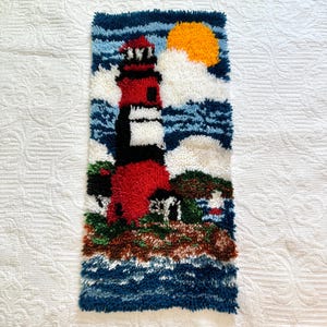 May include: A rectangular, handmade wall hanging featuring a lighthouse scene. The lighthouse is red and white, set against a blue sky with a yellow sun and white clouds. The ocean is depicted in shades of blue.