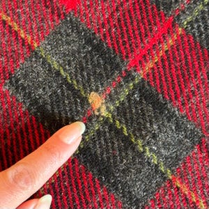 Puede incluir: Primer plano de una tela a cuadros roja, negra y verde. La tela tiene un aspecto texturizado con un patr&oacute;n repetitivo de l&iacute;neas rojas y formas de diamante negras. Un dedo apunta a la tela.