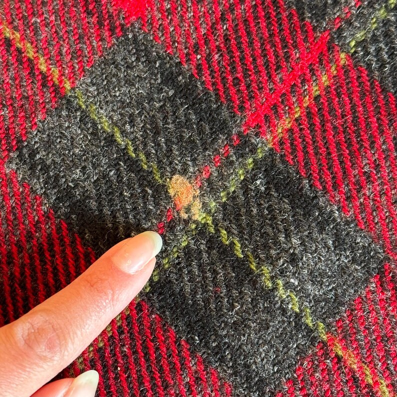 Puede incluir: Primer plano de una tela a cuadros roja, negra y verde. La tela tiene un aspecto texturizado con un patr&oacute;n repetitivo de l&iacute;neas rojas y formas de diamante negras. Un dedo apunta a la tela.