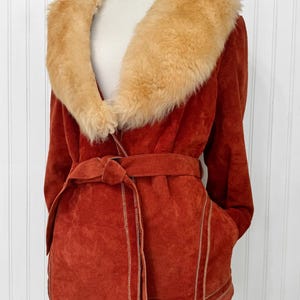 May include: Rust-colored suede jacket with a tan faux fur collar and a matching belt. The jacket features a wrap-style design with pockets and tan stitching details. A vintage-inspired outerwear piece.