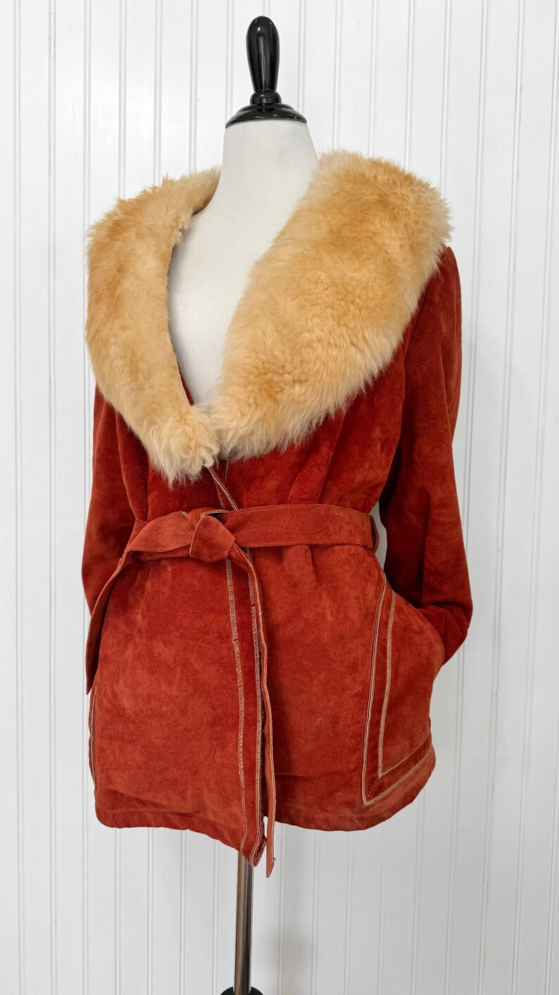May include: Rust-colored suede jacket with a tan faux fur collar and a matching belt. The jacket features a wrap-style design with pockets and tan stitching details. A vintage-inspired outerwear piece.