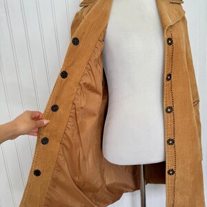 May include: A tan corduroy coat with a button-up front and a collared neckline. The coat features black buttons and decorative stitching. The interior lining is a lighter shade of tan. The coat is displayed on a mannequin.