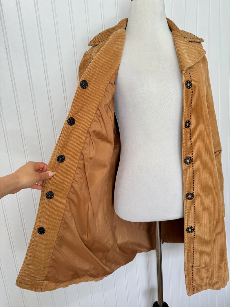 May include: A tan corduroy coat with a button-up front and a collared neckline. The coat features black buttons and decorative stitching. The interior lining is a lighter shade of tan. The coat is displayed on a mannequin.