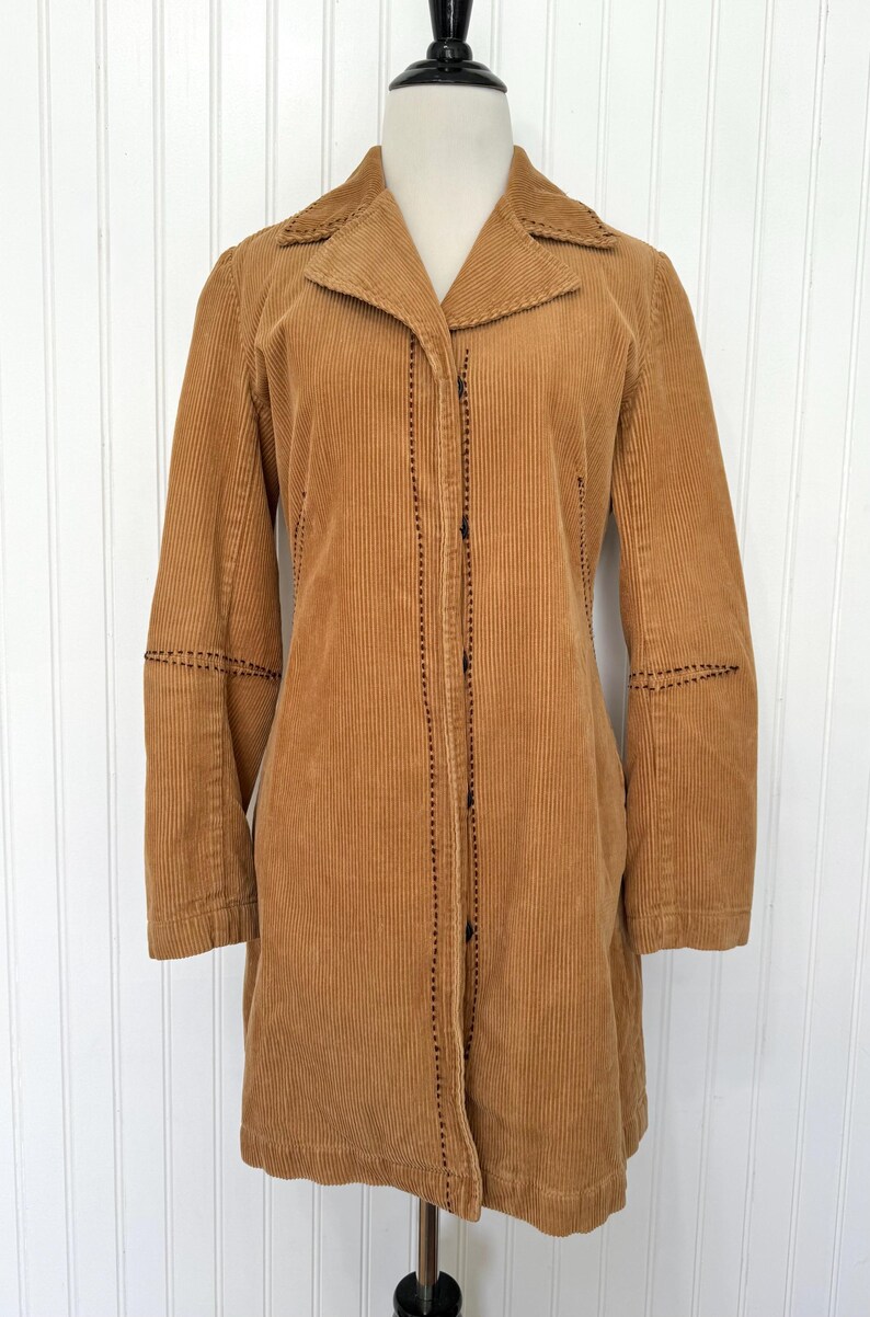 May include: A tan corduroy coat with a notched collar and long sleeves. The coat features black stitching along the seams and button closures down the front. The coat is displayed on a mannequin.