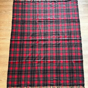 Puede incluir: Una manta rectangular con un estampado de cuadros rojos, negros y grises. La manta tiene flecos en los bordes. El dise&ntilde;o de cuadros presenta una cuadr&iacute;cula de cuadrados en los tres colores. La manta est&aacute; hecha de un material suave.