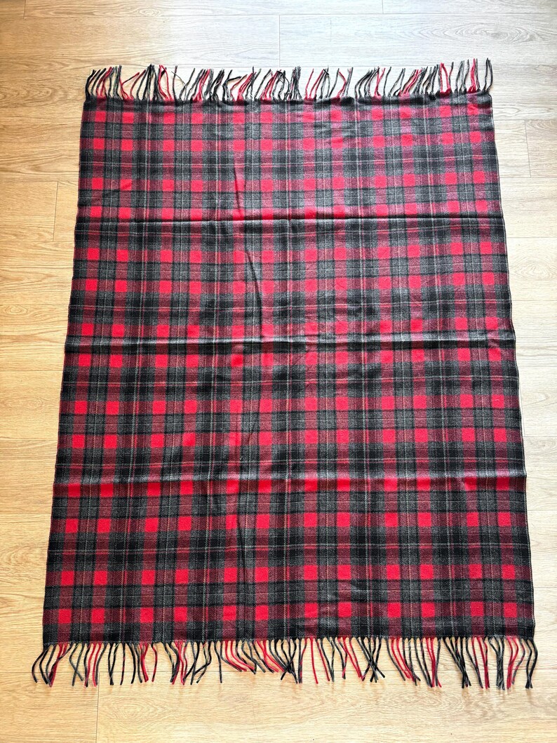 Puede incluir: Una manta rectangular con un estampado de cuadros rojos, negros y grises. La manta tiene flecos en los bordes. El dise&ntilde;o de cuadros presenta una cuadr&iacute;cula de cuadrados en los tres colores. La manta est&aacute; hecha de un material suave.