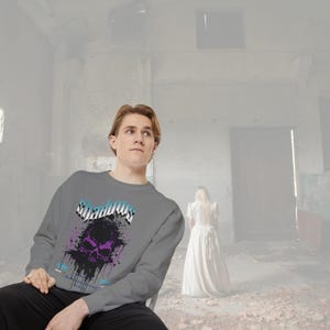 A man wearing a gray crewneck sweatshirt with a purple and black skull design, splattered with black ink, and the word "Shadows" above it in blue and white, sitting in a desolate room.
