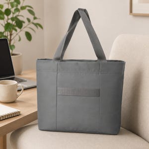A gray nylon tote bag with two shoulder straps sits upright on a beige upholstered chair next to a wooden desk with a laptop and mug.