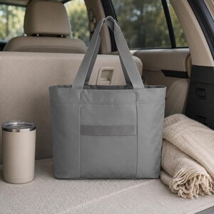 A plain gray tote bag is positioned in the back of a vehicle alongside a light-colored tumbler and a folded tan blanket.