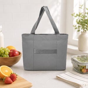 A plain gray tote bag sits on a kitchen counter next to a bowl of fruit, a cutting board with strawberries, a jar, and a container of salad.