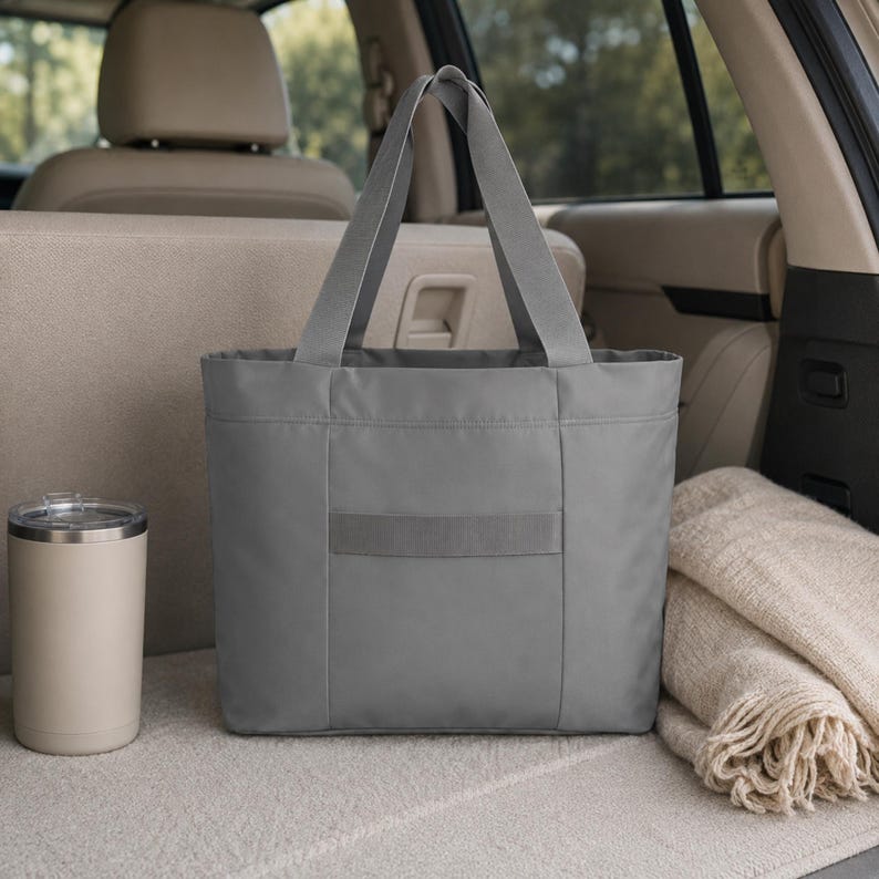 A gray tote bag is placed in the trunk of a car next to a tan tumbler and a folded blanket.