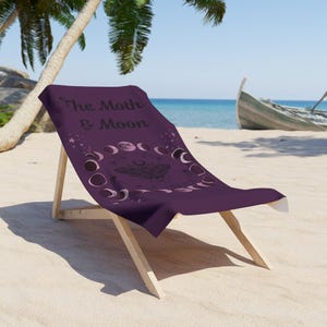 A purple beach towel with the text "The Moth & Moon" printed on it, sitting on a wooden chair on a sandy beach with palm trees and a boat visible in the background.