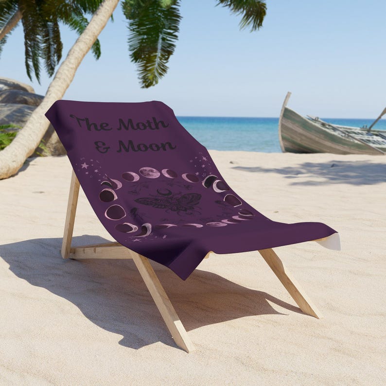 A purple beach towel with the text "The Moth & Moon" printed on it, sitting on a wooden chair on a sandy beach with palm trees and a boat visible in the background.