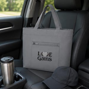 A gray tote bag sits on the black leather seat of a car next to a metal travel mug and a dark baseball cap.