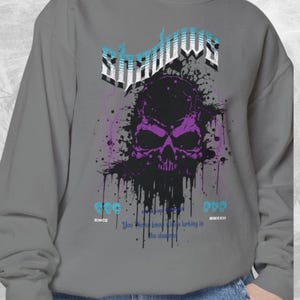 Close-up of a gray crewneck sweatshirt with a purple and black skull design, splattered with black ink, and the word "Shadows" above it in blue and white.