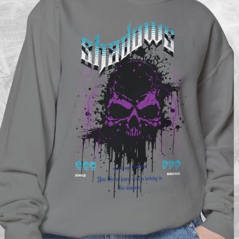 Close-up of a gray crewneck sweatshirt with a purple and black skull design, splattered with black ink, and the word "Shadows" above it in blue and white.