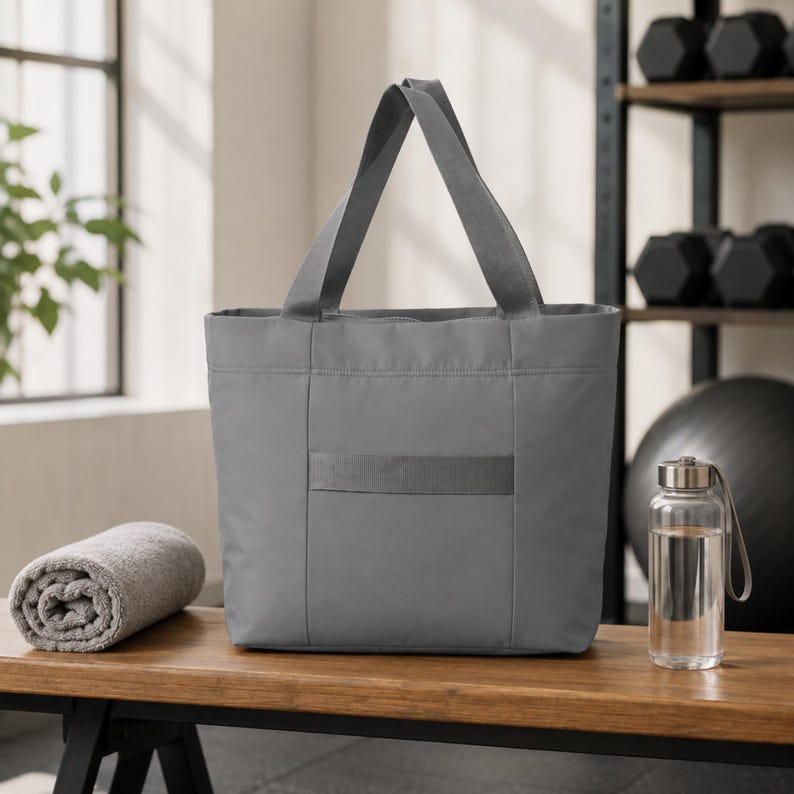 A gray tote bag sits on a wooden bench next to a rolled gray towel and a glass water bottle, with dumbbells and an exercise ball blurred in the background.