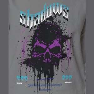 Close-up of a gray crewneck sweatshirt with a purple and black skull design, splattered with black ink, and the word "Shadows" above it in blue and white.