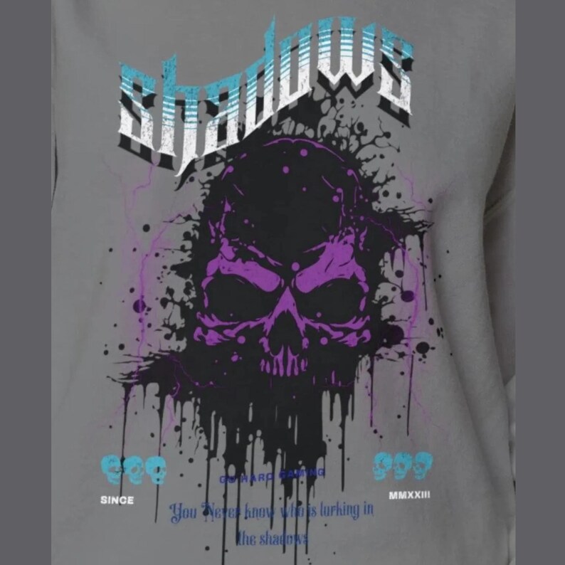 Close-up of a gray crewneck sweatshirt with a purple and black skull design, splattered with black ink, and the word "Shadows" above it in blue and white.