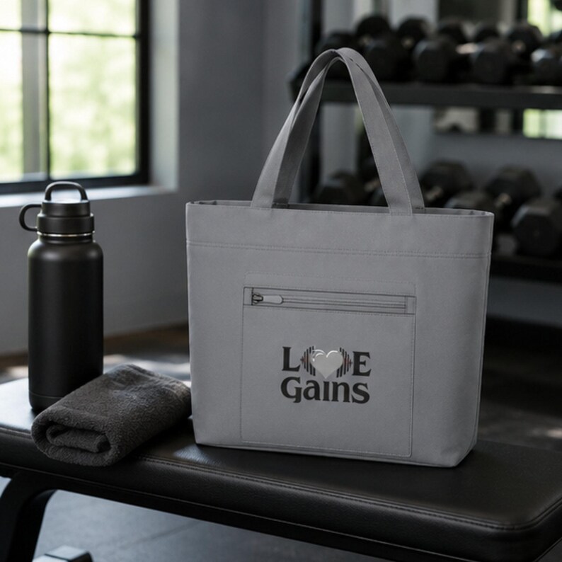 A gray tote bag with the Love Gains graphic rests on a gym bench alongside a black water bottle and a dark rolled towel.