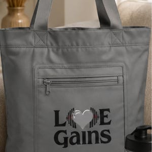 Close-up view of the gray tote bag featuring a zippered front pocket and the text Love Gains with a heart and dumbbell graphics.