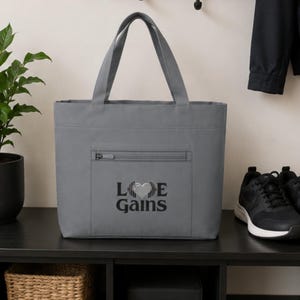 A gray tote bag with the Love Gains print on the front pocket sits on a black entryway table next to a pair of black sneakers.