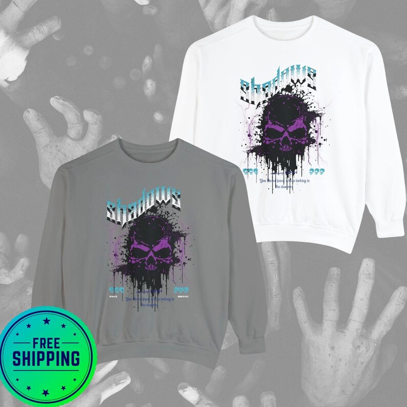 A gray crewneck sweatshirt and a white crewneck sweatshirt, both with a purple and black skull design, splattered with black ink, and the word "Shadows" above it in blue and white.