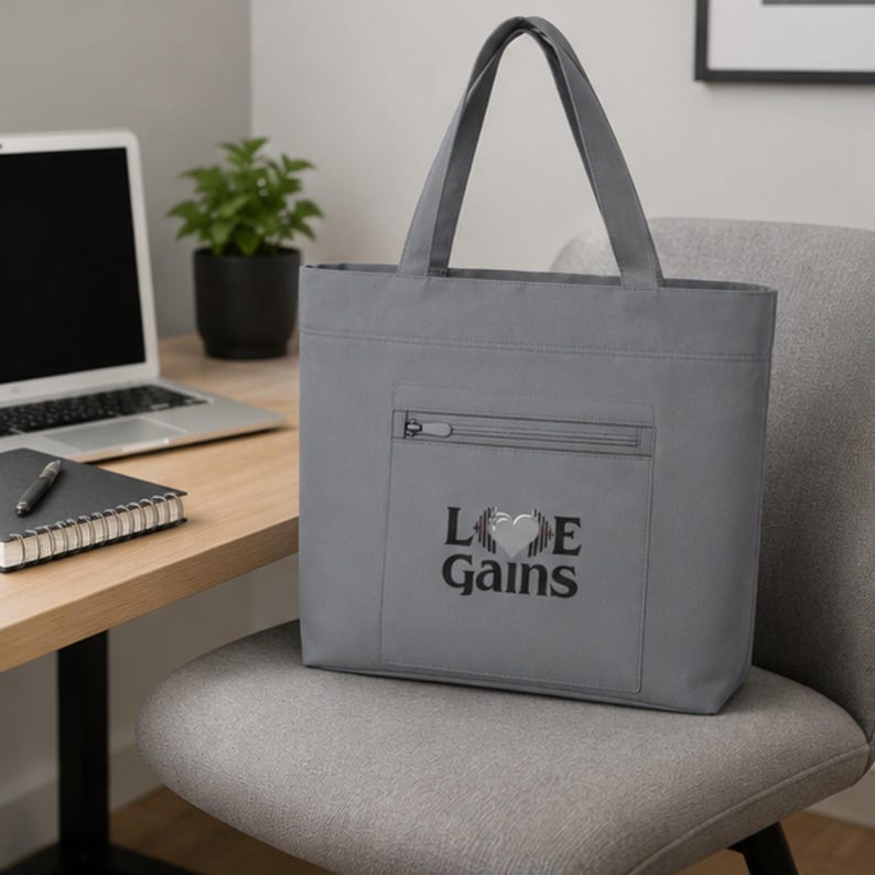 A gray tote bag with a zippered front pocket and Love Gains graphic sits on a gray office chair in front of a desk with a laptop.
