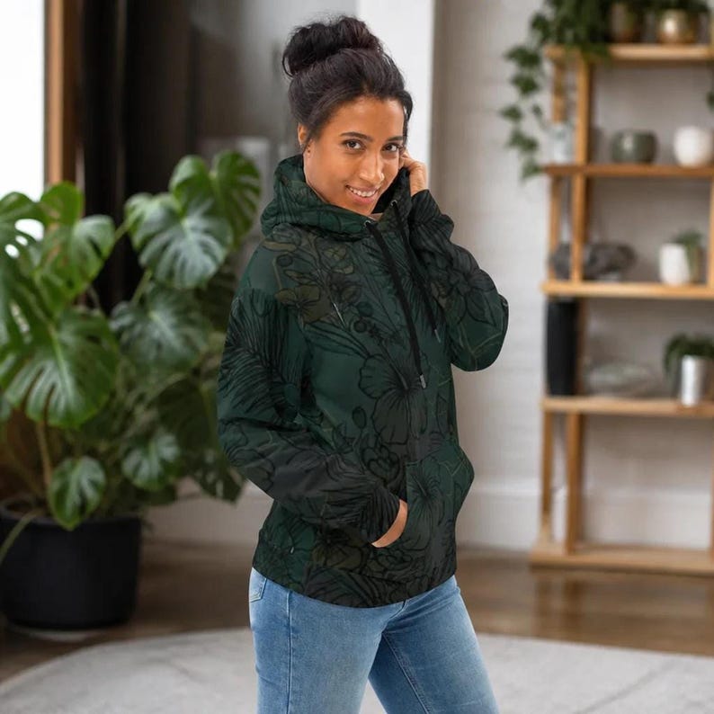 The woman turned slightly to the side in front of a potted plant, showing the profile of the dark green hoodie and its front pocket.