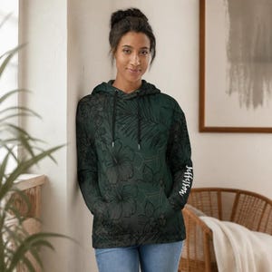 The same woman leaning against a wall indoors, wearing the dark green floral hoodie and blue jeans.