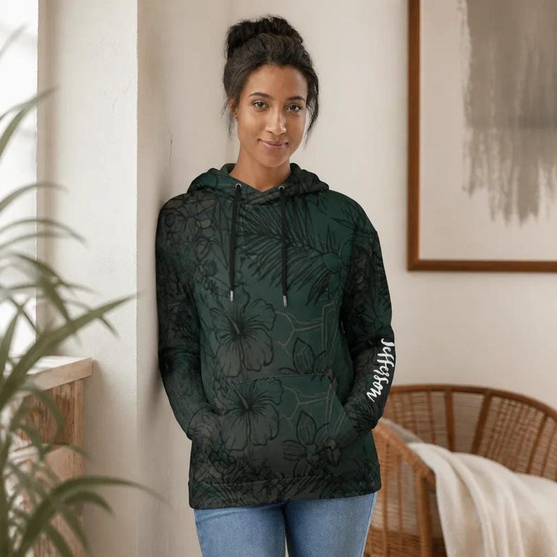 The same woman leaning against a wall indoors, wearing the dark green floral hoodie and blue jeans.