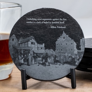 May include: A set of six black slate coasters with a medieval town scene engraved on the top. The coasters are in a black metal stand. The quote "Underlying most arguments against the free market is a lack of belief in freedom itself. - Milton Friedman" is engraved on the top coaster.