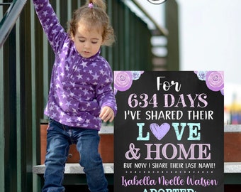 May include: A chalkboard sign with purple flowers and a pink heart announces that a child has been adopted. The sign reads "For 634 days I've shared their love & home, but now I share their last name! Isabella Noelle Watson Adopted: November 8, 2022". A young child in a purple star-patterned shirt and blue jeans stands on a set of stairs.