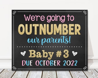 May include: Black chalkboard sign with gold glitter text announcing "We're going to OUTNUMBER our parents! Baby #3 DUE OCTOBER 2022".