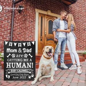 Könnte beinhalten: Ein schwarzes Schild aus Schiefer mit weißem Text, das die Ankunft eines neuen Menschen ankündigt. Das Schild lautet "Mama und Papa bekommen mir einen Menschen! Baby Caldwell Wachhund-Dienst beginnt Juni 2022". Ein Paar steht vor dem Schild, und ein Golden Retriever sitzt vor ihnen.