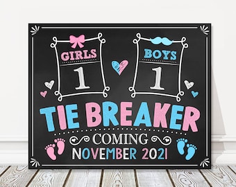 May include: A black chalkboard sign with the text "Girls 1  Boys 1 Tie Breaker Coming November 2021" in pink and blue. The sign has a pink bow, a blue mustache, and footprints.