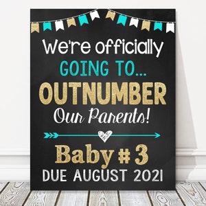 May include: A black chalkboard sign with white and gold lettering announces a pregnancy with the text "We're officially GOING TO... OUTNUMBER Our Parents! Baby #3 DUE AUGUST 2021". The sign is decorated with turquoise and white bunting.