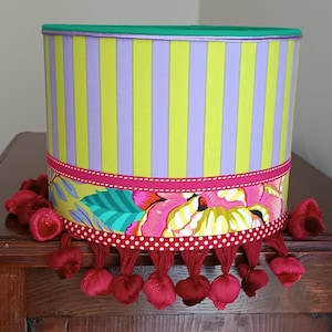 May include: A cylindrical lampshade with vertical stripes in yellow and lilac. A floral patterned band with pink, red, and green accents encircles the shade. Red pom-poms hang from the bottom edge. The top edge is green.