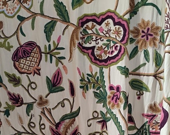 May include: Cream-colored fabric with intricate floral embroidery. The design features large, stylized flowers in shades of pink, purple, and brown, with green leaves and stems. The embroidery is detailed and creates a textured effect.
