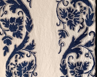 May include: White fabric with two vertical borders of embroidered blue floral and leaf patterns. The embroidery has a detailed design with a gray outline. The fabric is likely used for home decor.