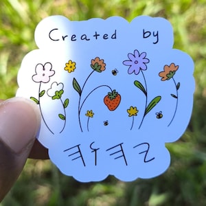 Peut inclure: Un autocollant blanc avec un motif floral et les mots "Created by" en haut. L'autocollant présente diverses fleurs, une fraise et du texte en hébreu. L'autocollant est tenu sur un fond d'herbe verte.