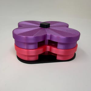 Pode incluir: Um recipiente multicamadas em forma de flor em tons de roxo e rosa, com uma base preta e um pequeno quadrado preto no topo. O recipiente &eacute; feito de pl&aacute;stico e tem uma superf&iacute;cie texturizada.