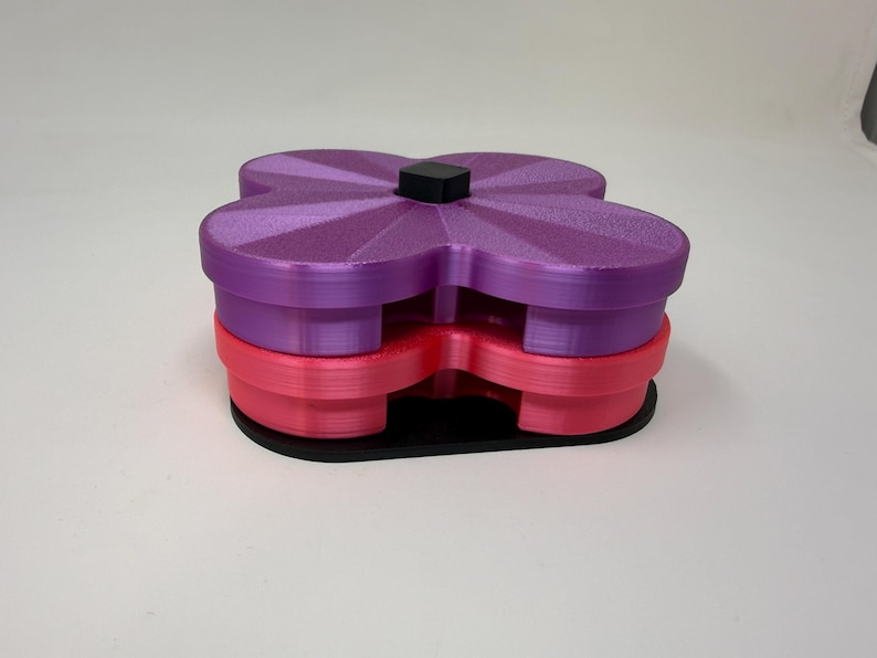 Pode incluir: Um recipiente multicamadas em forma de flor em tons de roxo e rosa, com uma base preta e um pequeno quadrado preto no topo. O recipiente &eacute; feito de pl&aacute;stico e tem uma superf&iacute;cie texturizada.