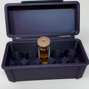 Puede incluir: Una caja rectangular de color púrpura oscuro con tapa abierta, que contiene un pequeño vial de vidrio con líquido marrón. La caja tiene cuatro compartimentos moldeados y una tapa con bisagras. El vial tiene una tapa dorada.