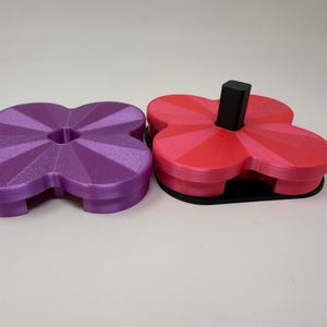 Pode incluir: Dois objetos impressos em 3D em forma de trevo. Um &eacute; roxo, o outro &eacute; vermelho e rosa com um poste retangular preto. Ambos t&ecirc;m uma superf&iacute;cie texturizada e est&atilde;o em um fundo branco. Os objetos parecem ser porta-copos ou pequenas bandejas.