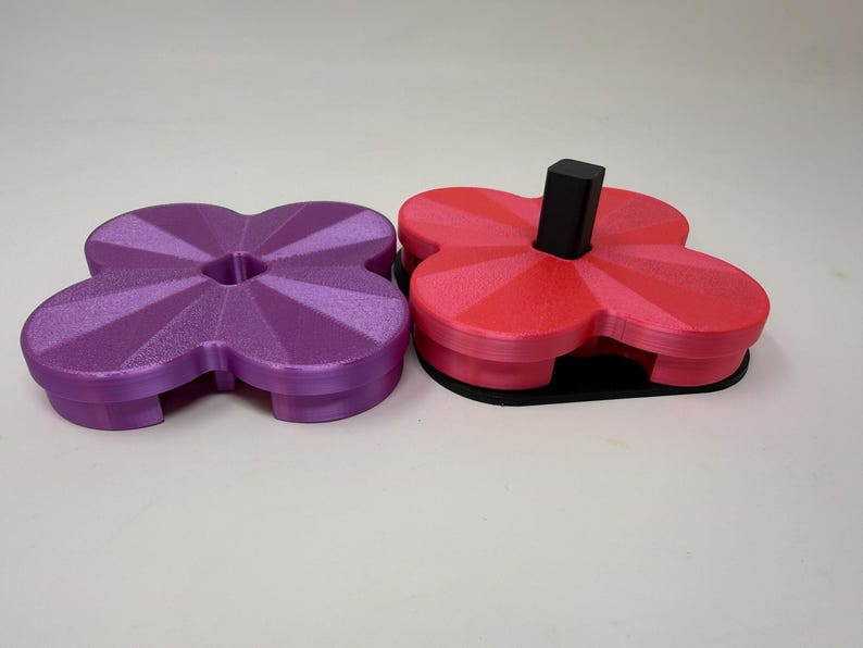 Pode incluir: Dois objetos impressos em 3D em forma de trevo. Um &eacute; roxo, o outro &eacute; vermelho e rosa com um poste retangular preto. Ambos t&ecirc;m uma superf&iacute;cie texturizada e est&atilde;o em um fundo branco. Os objetos parecem ser porta-copos ou pequenas bandejas.