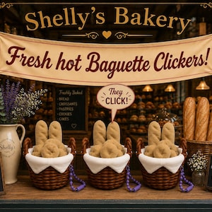 Op de afbeelding: Een etalage van een bakkerij met een banner Shelly's Bakery Fresh hot Baguette Clickers! Drie bruine rieten manden bevatten pluche brood, elk met een paars kralenkoord. Een tekstballon zegt THEY CLICK! Kleine krijtborden en een vaas met lavendel zijn aanwezig.