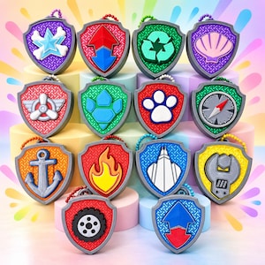 May include: A collection of fourteen shield-shaped plastic badges featuring various embossed icons including a star, recycling symbol, seashell, propeller, paw print, compass, anchor, flame, building, wrench, and tire, each with a small colored chain.